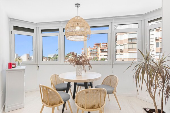 A room with a round table, six wicker chairs, a hanging light fixture, and a potted plant.