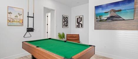 Una mesa de billar en una habitación con paredes blancas, una puerta y obras de arte enmarcadas.