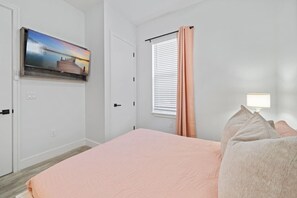 A bedroom with a bed, a TV mounted on the wall, a window with blinds, and a bedside lamp. - The Palms at Vincente (Panama City Beach)