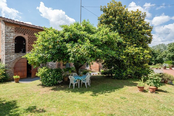 Un edificio de ladrillo con un gran árbol en el patio, una mesa con sillas y plantas en macetas.