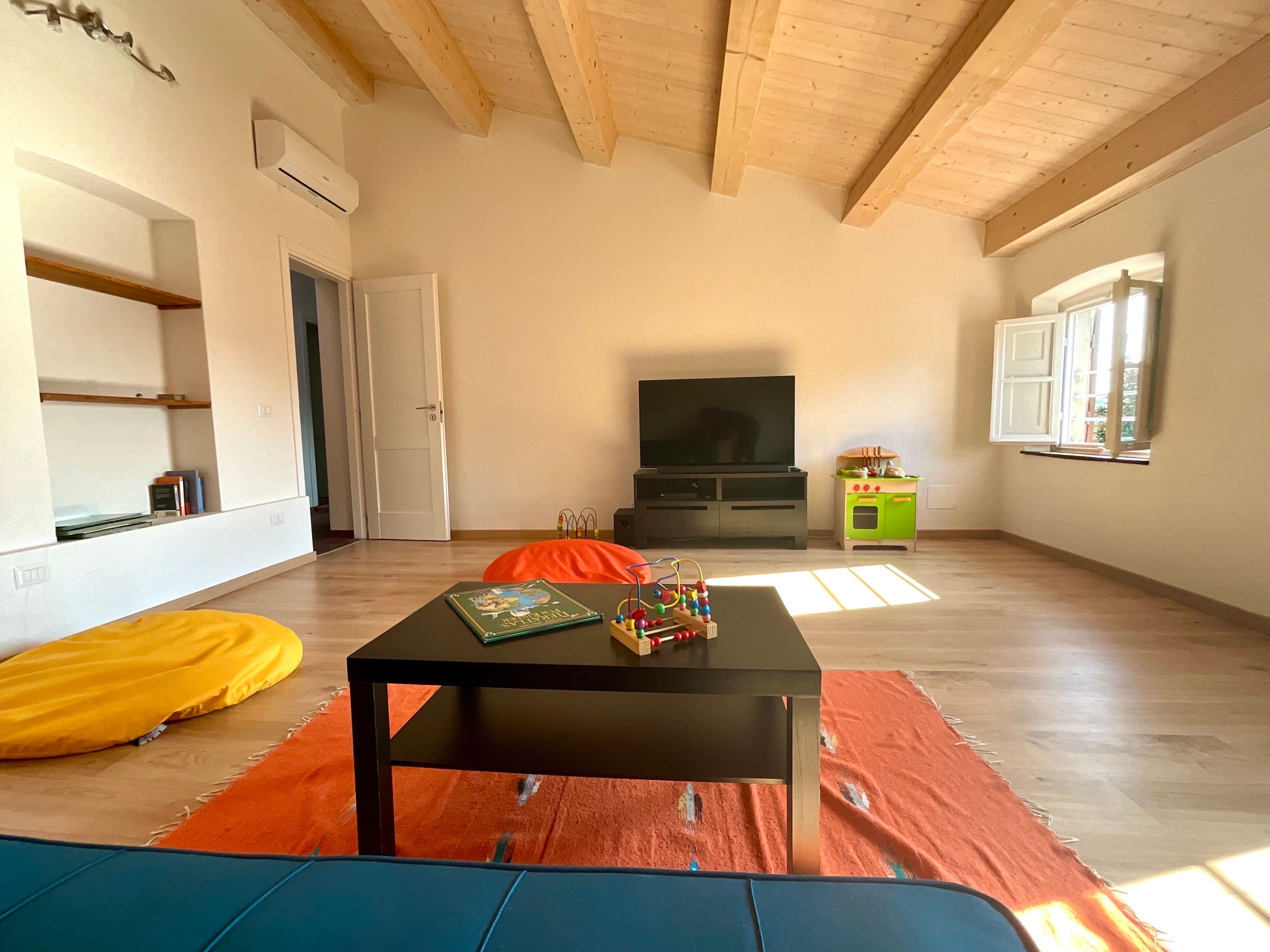 Een ruime woonkamer met houten plafond, een flatscreen televisie, een kleine keukenhoek en een bank.