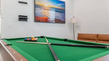 Ein Billardtisch mit Kugeln und Spielstöcken, eine Couch, eine Lampe und ein Gemälde mit Sonnenuntergang über dem Meer.
