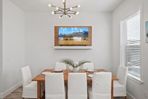 A dining area with a wooden table, white chairs, a modern chandelier, and a framed landscape painting on the wall.