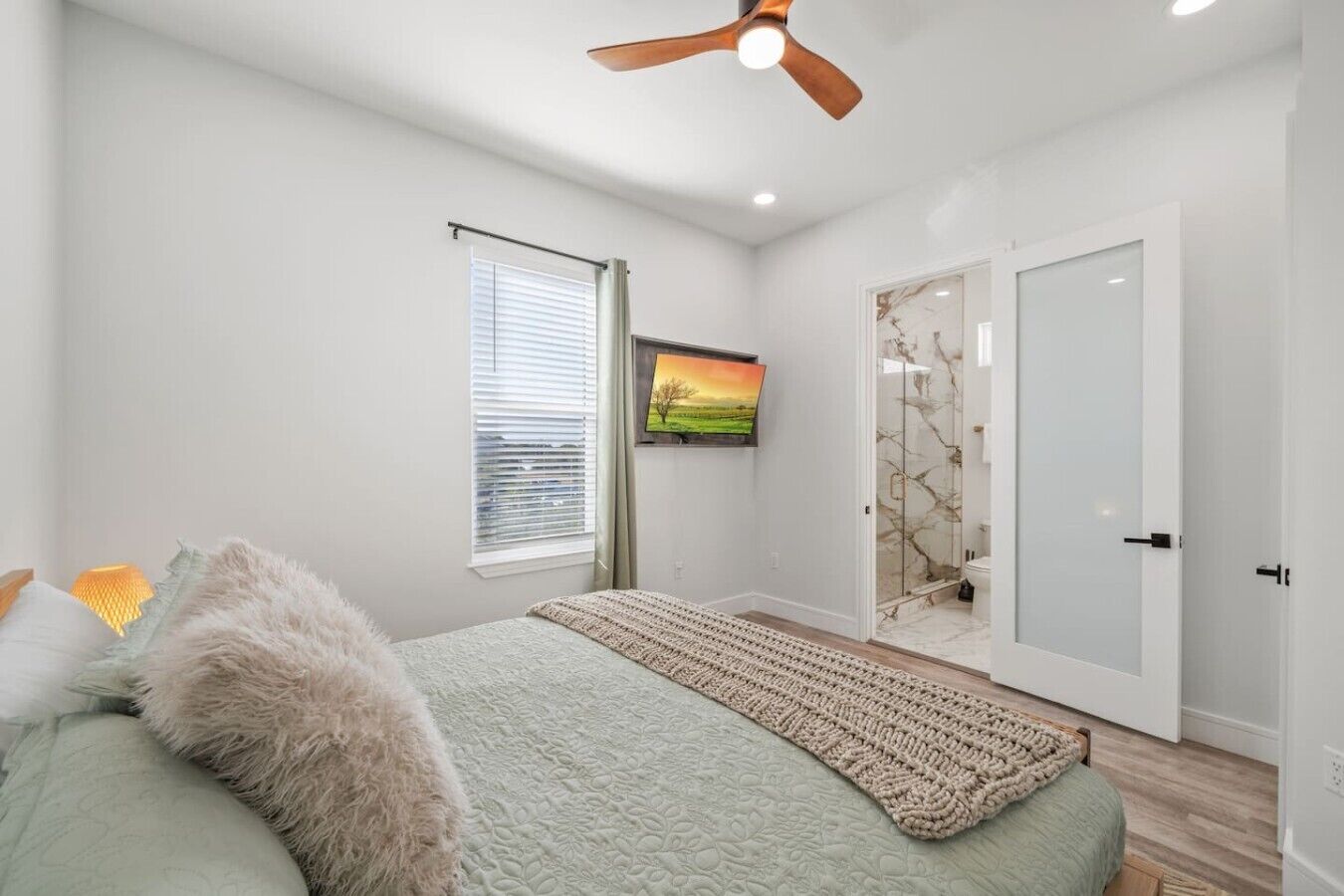A bedroom with a bed, a ceiling fan, a window with blinds, a painting, and a door with a glass panel.