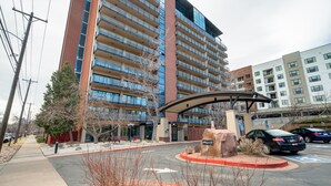 A multi-story building with a modern architectural design, featuring a mix of glass and brick facades, and a parking area with marked spaces. - Downtown City Walk High-Rise 2BR | GYM | Sauna (Colorado Springs)