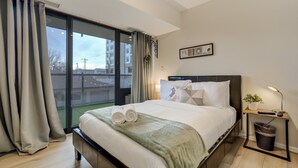 A modern hotel room with a large bed, a bedside table with a lamp, a small plant, and a view of the outside through a large window. - Downtown City Walk High-Rise 2BR | GYM | Sauna (Colorado Springs)