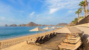 Une plage bordée de rangées de transats et de parasols, un rivage sablonneux et une mer d’un bleu limpide, avec des montagnes qui se dessinent à l’horizon.