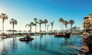 A resort pool area with lounge chairs and umbrellas, surrounded by palm trees and a mountain backdrop. - Beachfront 5 Star Luxury Resort, 2BR 3 Bath   July26-August 2 (Cabo San Lucas)