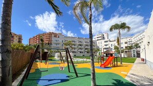 Um parquinho com balanços e um escorregador, cercado por palmeiras e edifícios.