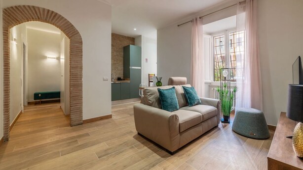 Una sala moderna con un sofá, una mesa de centro y una televisión. Se aprecia una cocina al fondo, un balcón con plantas y un arco de ladrillo que conduce a otra habitación.