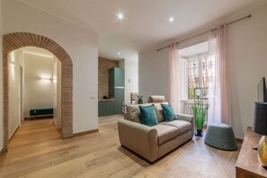 Een moderne woonkamer met een bank, een salontafel en een televisie. Achtergrond is een keuken, een balkon met planten, en een bakstenen boog die naar een andere kamer leidt.