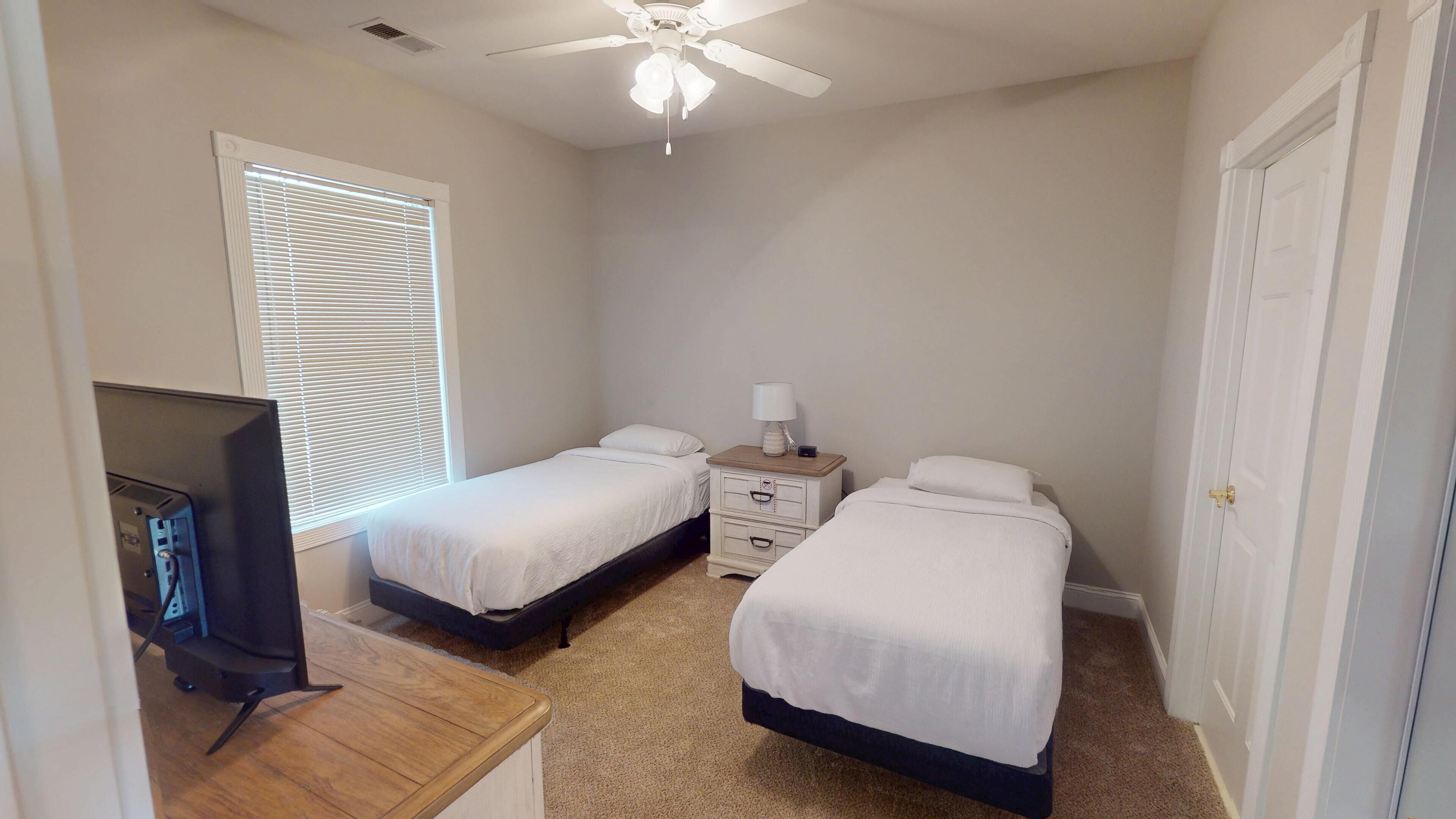 Une chambre avec deux lits simples, une table de chevet avec une lampe, un ventilateur de plafond et un téléviseur posé sur un meuble en bois.