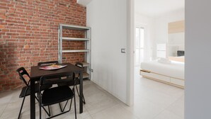 Une pièce avec un mur en briques, une porte blanche donnant sur une autre pièce, un lit avec un matelas blanc et une table avec des chaises.