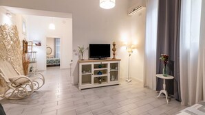 Un soggiorno moderno con una televisione su un mobile bianco, una poltrona a dondolo, una lampada da terra e un tavolino con un vaso.