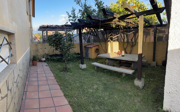 En uteplats med en gångväg av plattor, en pergola av trä, ett bord med en vas och krukväxter.