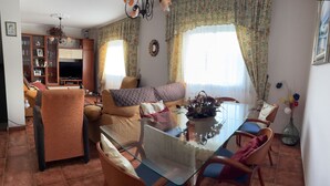 Una sala de estar con un sofá, una mesa de comedor de vidrio y sillas. Se ve un televisor y una estantería al fondo.