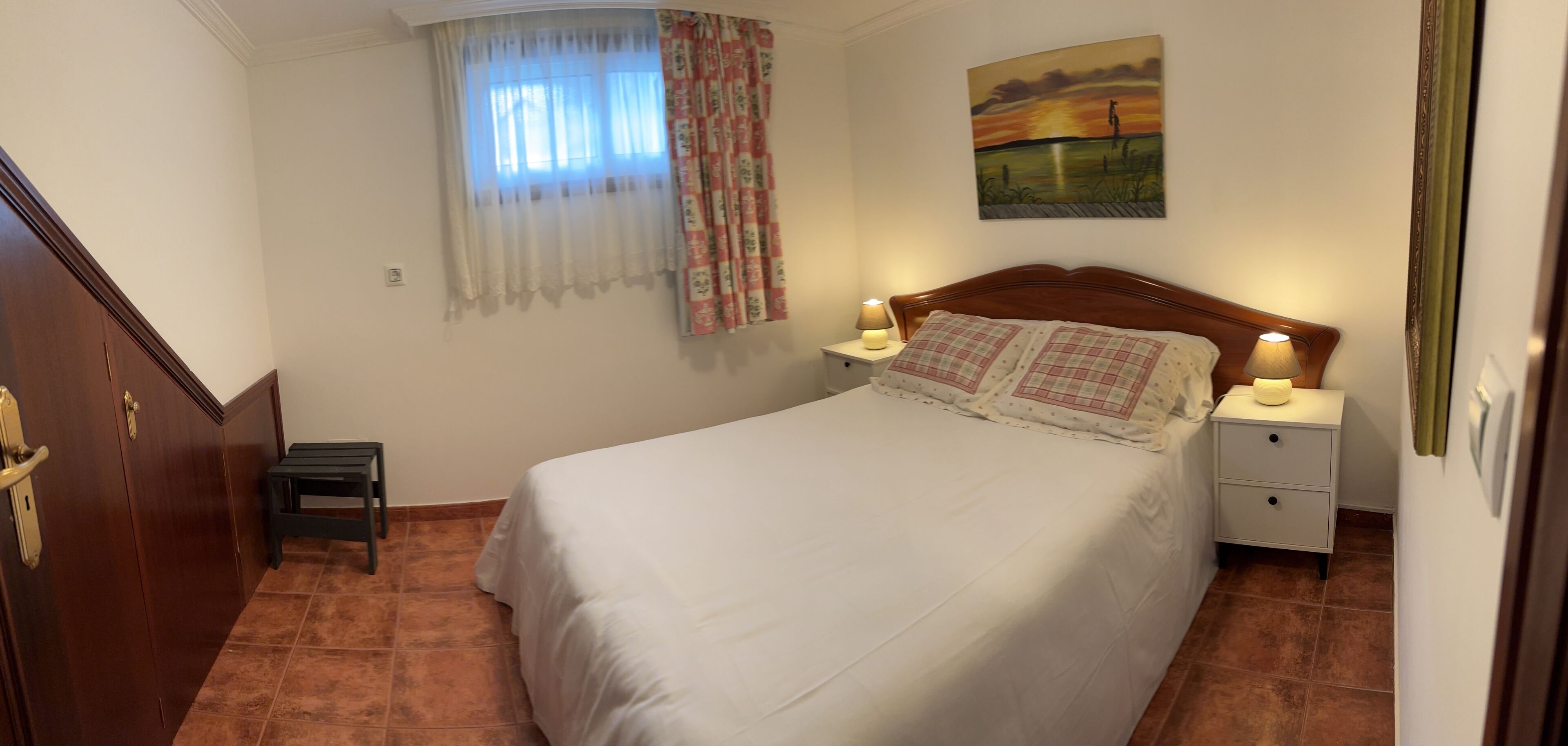 Un dormitorio con cama, mesitas de noche, lámpara, una pequeña mesa, ventana con cortinas y un cuadro en la pared.