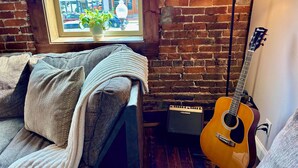 Ett mysigt rum med en tegelvägg, ett fönster med utsikt över byggnader, en soffa med en filt, en gitarr och en högtalare.