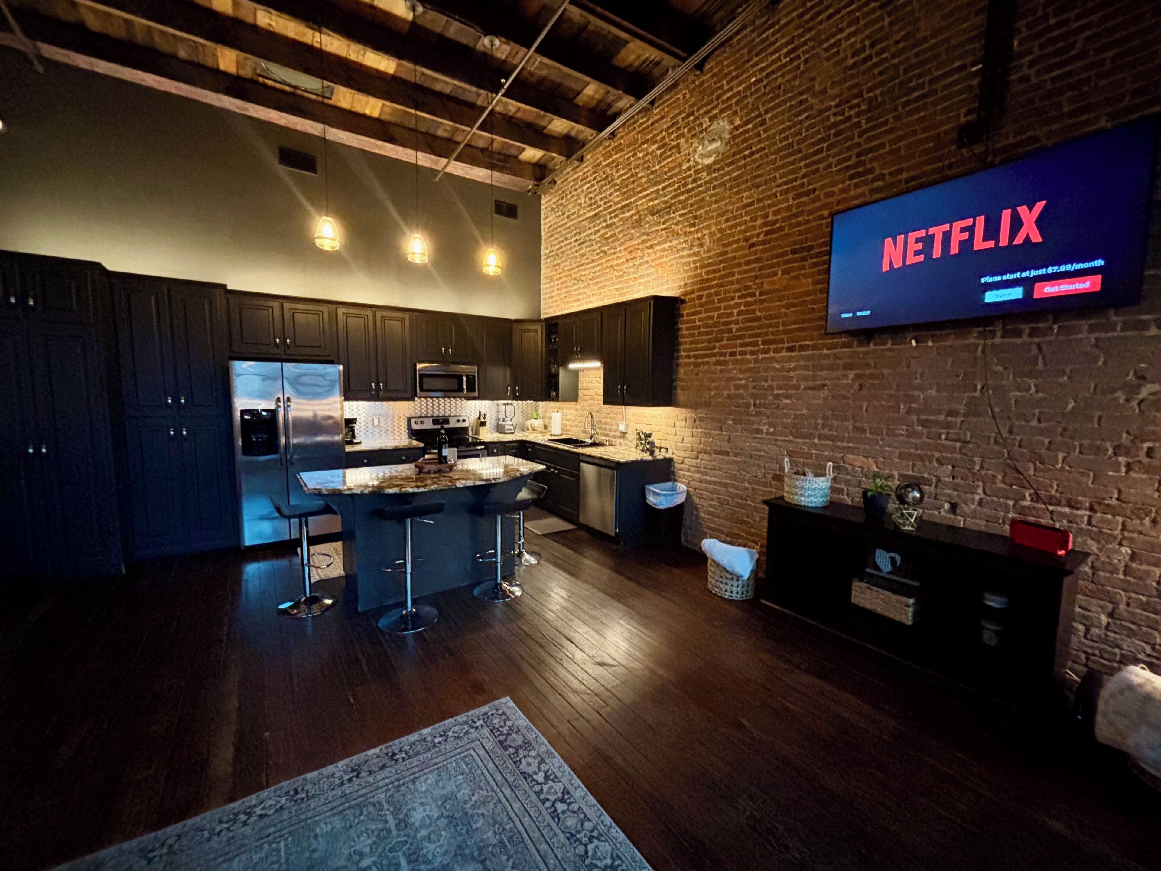 A modern kitchen with dark cabinetry, a central island with bar stools, a brick wall, and a large flat-screen TV displaying 'NETFLIX'.