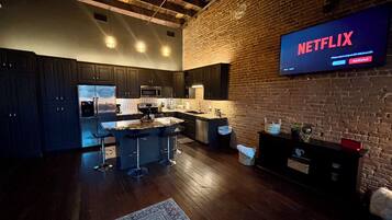 A modern kitchen with dark cabinetry, a central island with bar stools, a brick wall, and a large flat-screen TV displaying 'NETFLIX'.