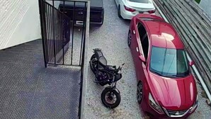 En parkeringsplats med bilar och en motorcykel, en grind vid ingången och en byggnad med en vit fasad.
