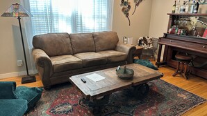 Una sala de estar con un sofá marrón, una mesa de centro de madera, un tapete estampado, un piano y una lámpara.