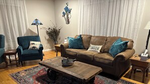 Una sala de estar con sofá marrón, sillón azul, mesa de centro de madera y alfombra estampada.