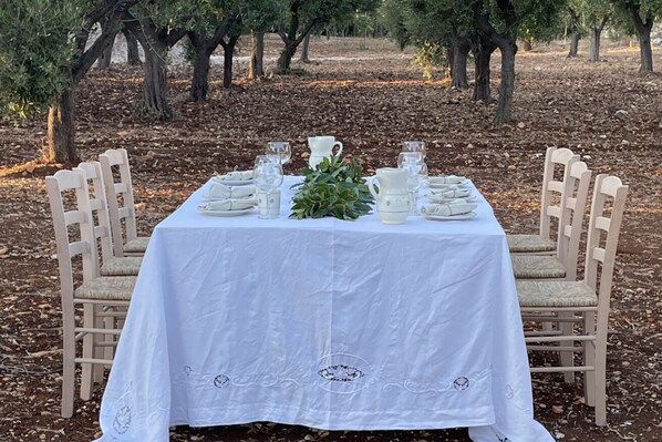 Une table d’extérieur, recouverte d’une nappe blanche et entourée de chaises, se trouve au milieu des arbres.