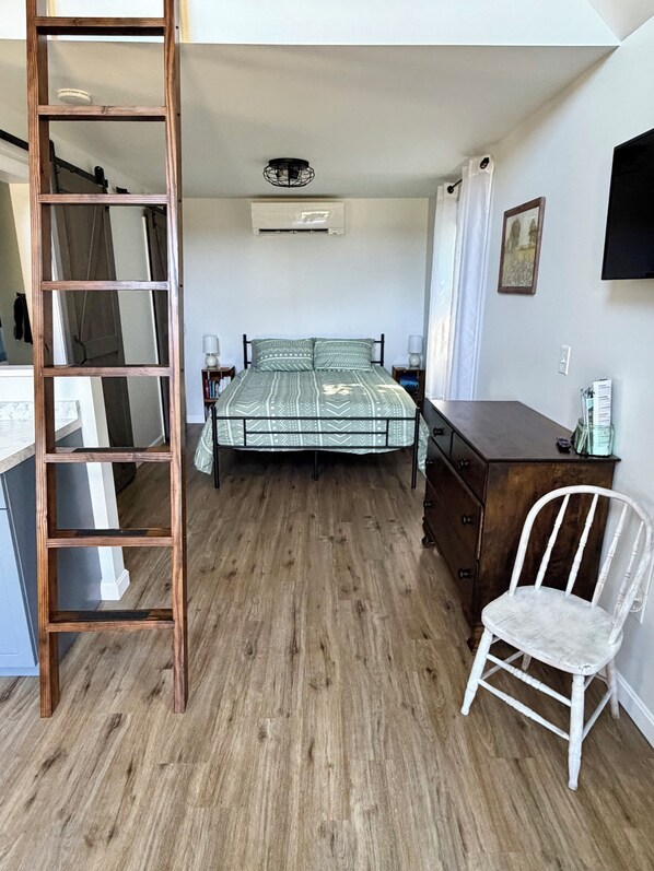 Une chambre avec une échelle en bois, un lit avec une literie à motifs, une chaise blanche, une commode et un téléviseur à écran plat fixé au mur.