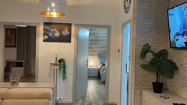 Ein modernes Wohnzimmer mit einem beigen Sofa, einer großen Pendelleuchte, einem Flachbildfernseher an einer Ziegelwand und einem Topf mit Pflanze auf einem Schrank.