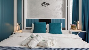 A hotel room with a bed, two pillows, a headboard, a nightstand, a lamp, a plant, and a wall decoration.