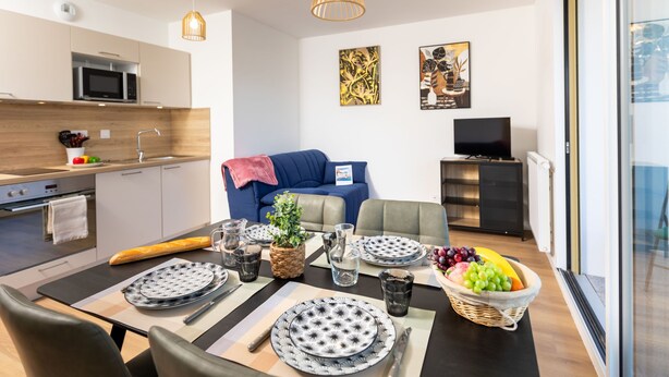Une cuisine moderne équipée d’un four à micro-ondes, d’une cuisinière et d’un évier. On y trouve également un coin repas avec une table dressée pour deux personnes, des assiettes et des verres. Un canapé bleu et un téléviseur se trouvent dans l’espace salon.