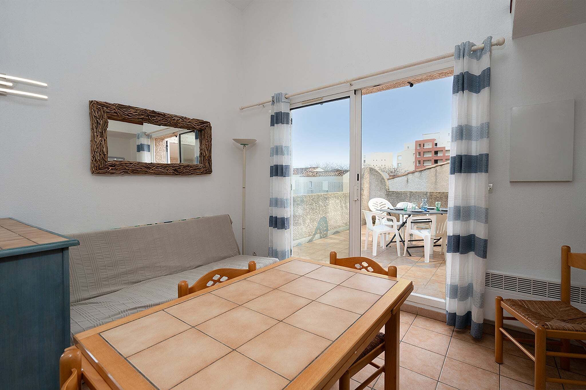 Ein Zimmer mit Holztisch, einer Couch, einem Spiegel und Blick auf einen Balkon mit Stühlen und Tisch.
