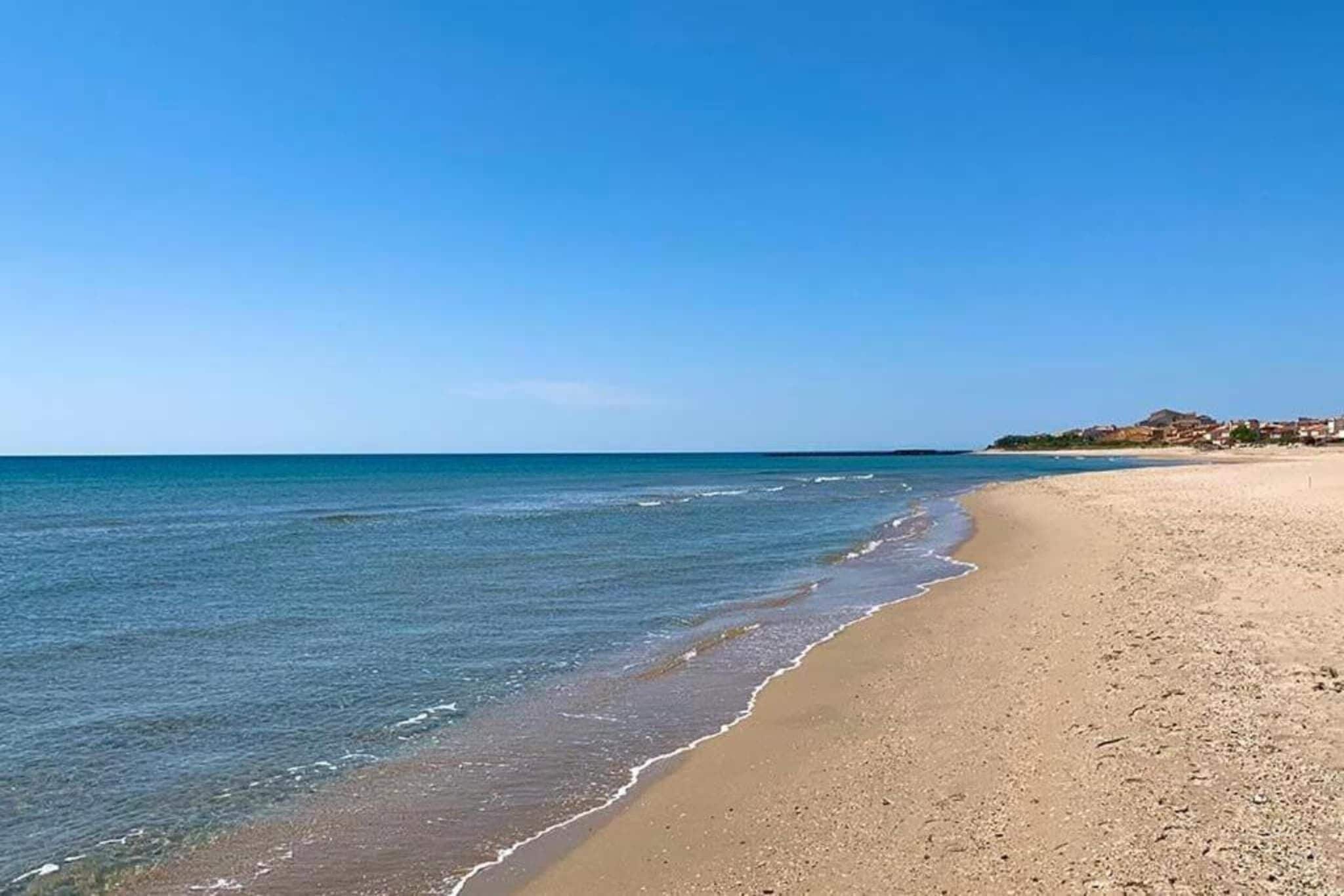 Une plage à l’eau d’un bleu limpide, bordée d’un rivage sablonneux, avec une ville qui se devine au loin.