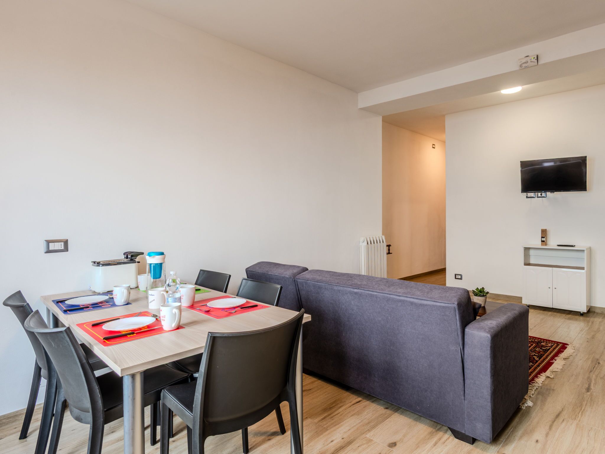 A modern living room with a dining area, a grey sofa, a flat-screen TV mounted on the wall, and a radiator.