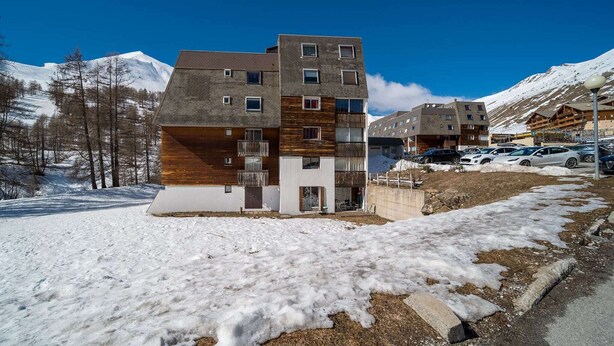 Edifici modern de diverses plantes amb façanes de fusta i formigó, ubicat en una zona muntanyosa nevada amb cel blau clar.
