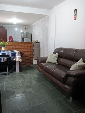 A living room with a brown leather sofa, a dining table with a patterned tablecloth, and a kitchen area with a brick wall and tiled backsplash.
