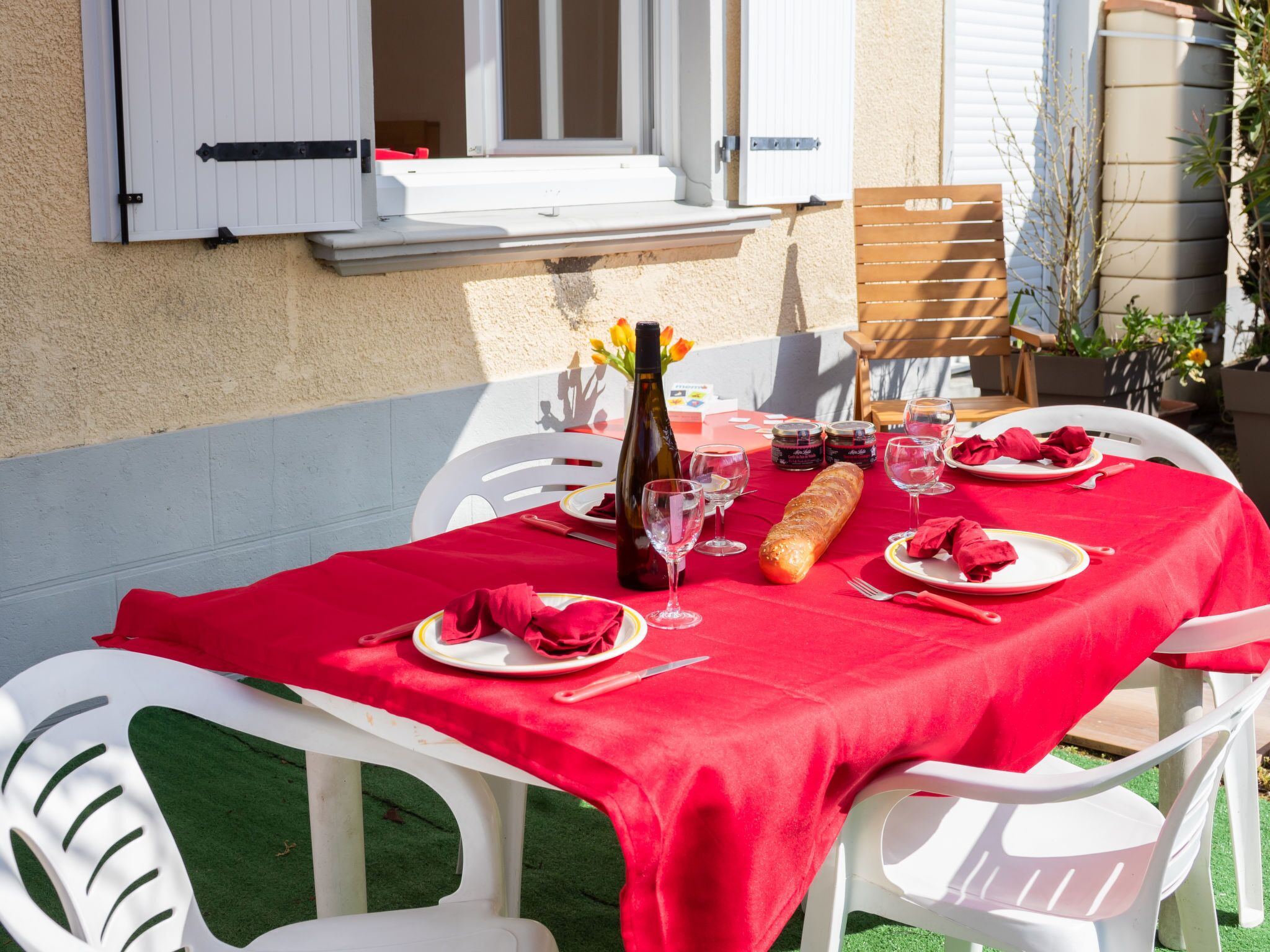 An outdoor dining area with a red tablecloth, white chairs, and a bottle of wine.