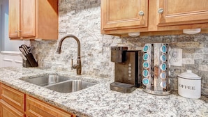 A kitchen with granite countertops, a stainless steel sink, wooden cabinets, a coffee maker, and a coffee storage container.