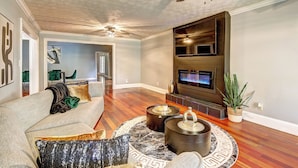 A spacious living room with a fireplace, a ceiling fan, and a flat-screen TV mounted on the wall.