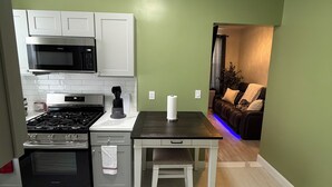 Cocina compacta con armarios blancos, microondas, estufa y una pequeña mesa con un banco.