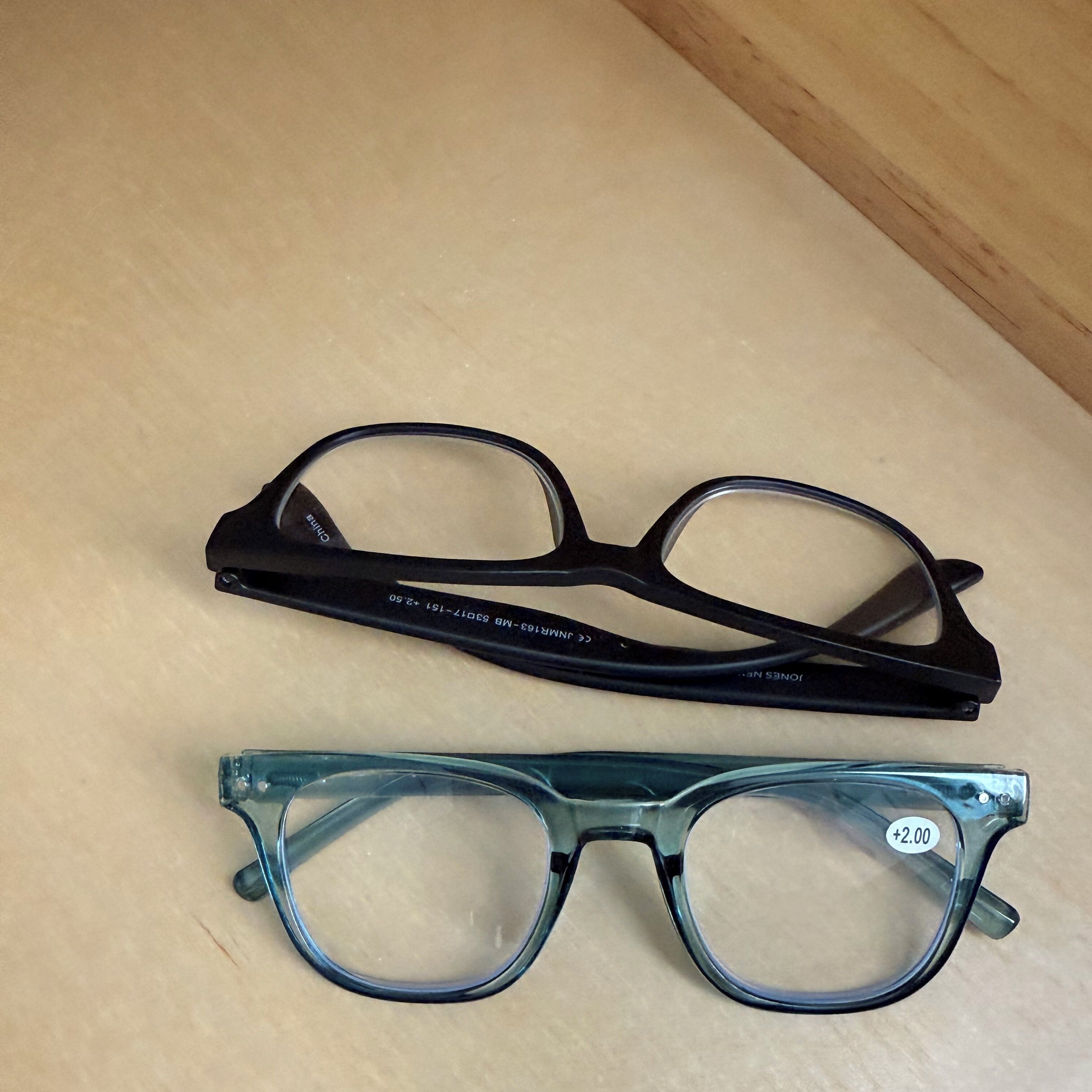 A pair of eyeglasses with a black frame and a pair with a clear frame, both placed on a wooden surface.