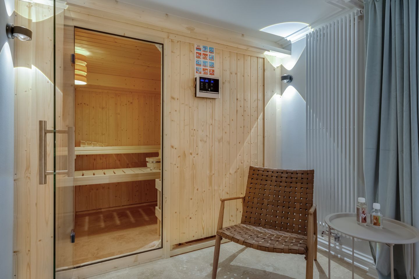 A sauna room with wooden walls, a glass door, a control panel, a woven chair, and a small table with bottles.