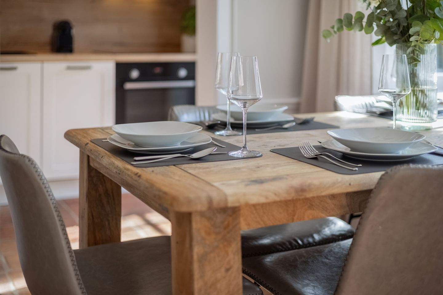 Una mesa de comedor dispuesta con platos, cubiertos y copas de vino, con una cocina al fondo.