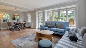 Ein geräumiges Wohnzimmer mit einem blauen Sofa, einem Esstisch mit Stühlen und großen Fenstern mit Blick auf Grünflächen.