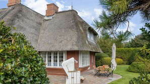 Ein reetgedecktes Cottage mit einer Steinterrasse, weißen Korbmöbeln und einem gepflegten Garten.