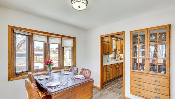 Een eetruimte met een houten tafel en stoelen, een vaas met bloemen, en een keuken met houten keukenkastjes en een inbouwkast met glazen deuren.