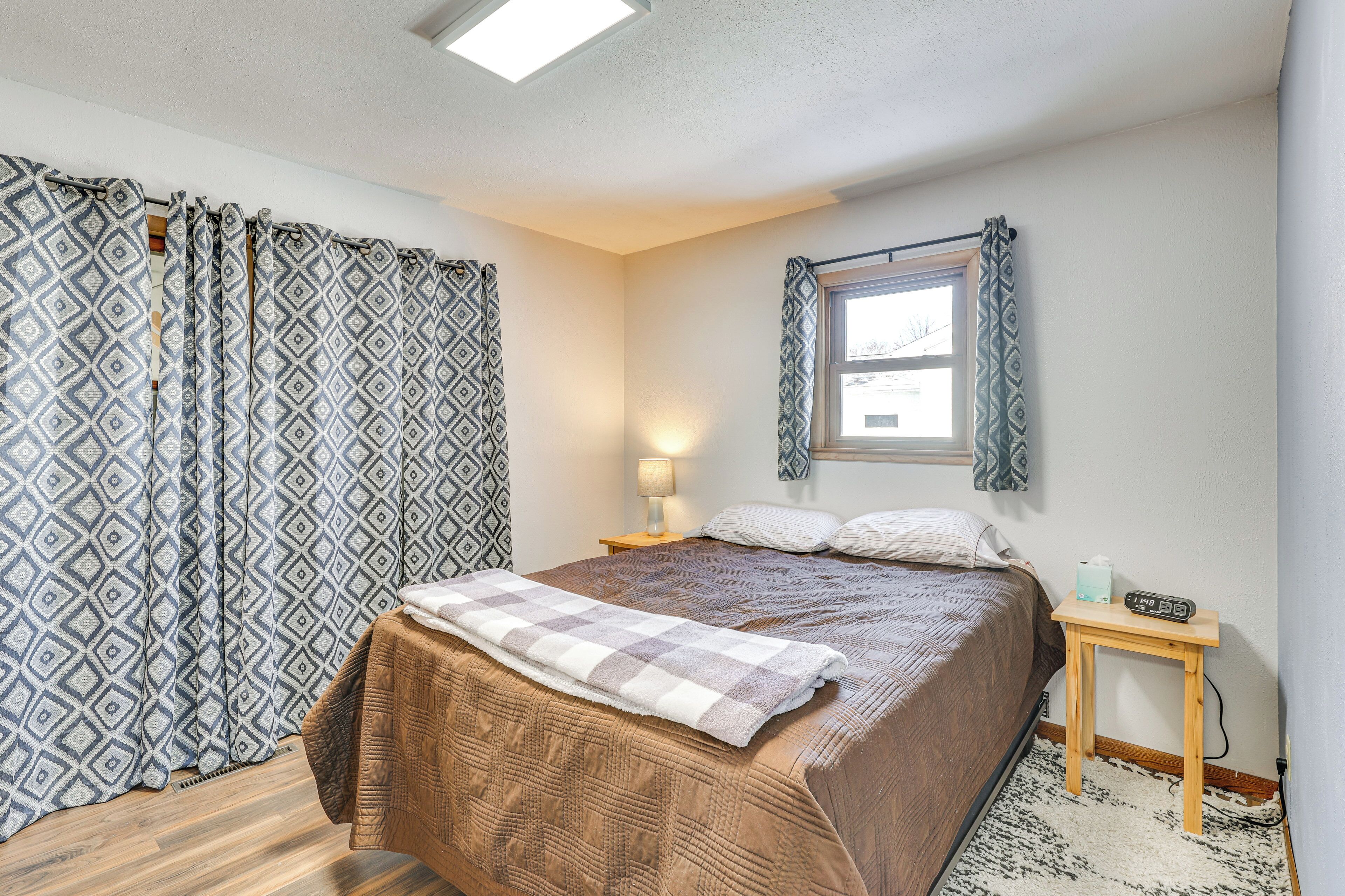 A bedroom with a large bed, patterned curtains, a bedside table with a lamp, and a window with a view.