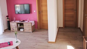 A modern living room with a pink wall, wooden flooring, a TV mounted on the wall, a wooden door, and a white table with a vase and remote control. - Villa Silvia by Interhome (Makarska)