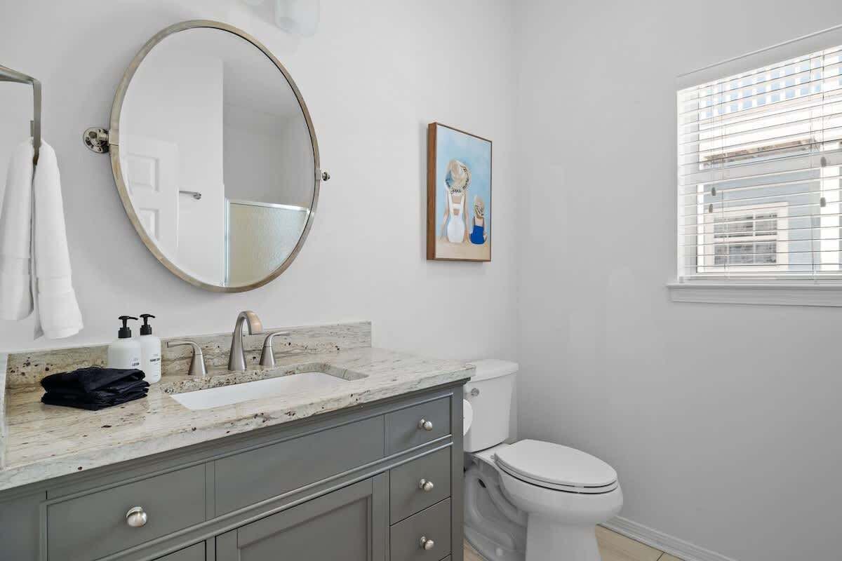 A bathroom with a round mirror, a sink with a marble countertop, a toilet, and a window with blinds.